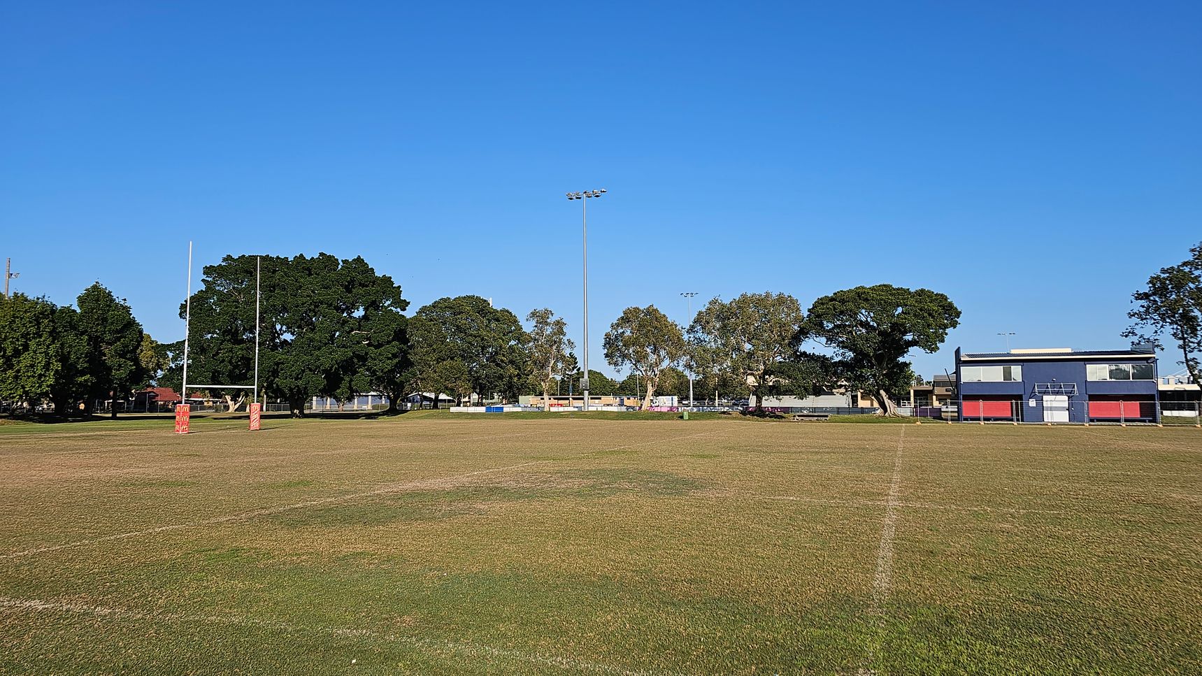 A rugby field with goalposts on the left, featuring red padding. The grass is patchy and worn. In the background, there are several large trees and a tall floodlight. On the right, a blue and red building with large windows is visible. The scene is open and spacious, with a clear view of the surrounding area.