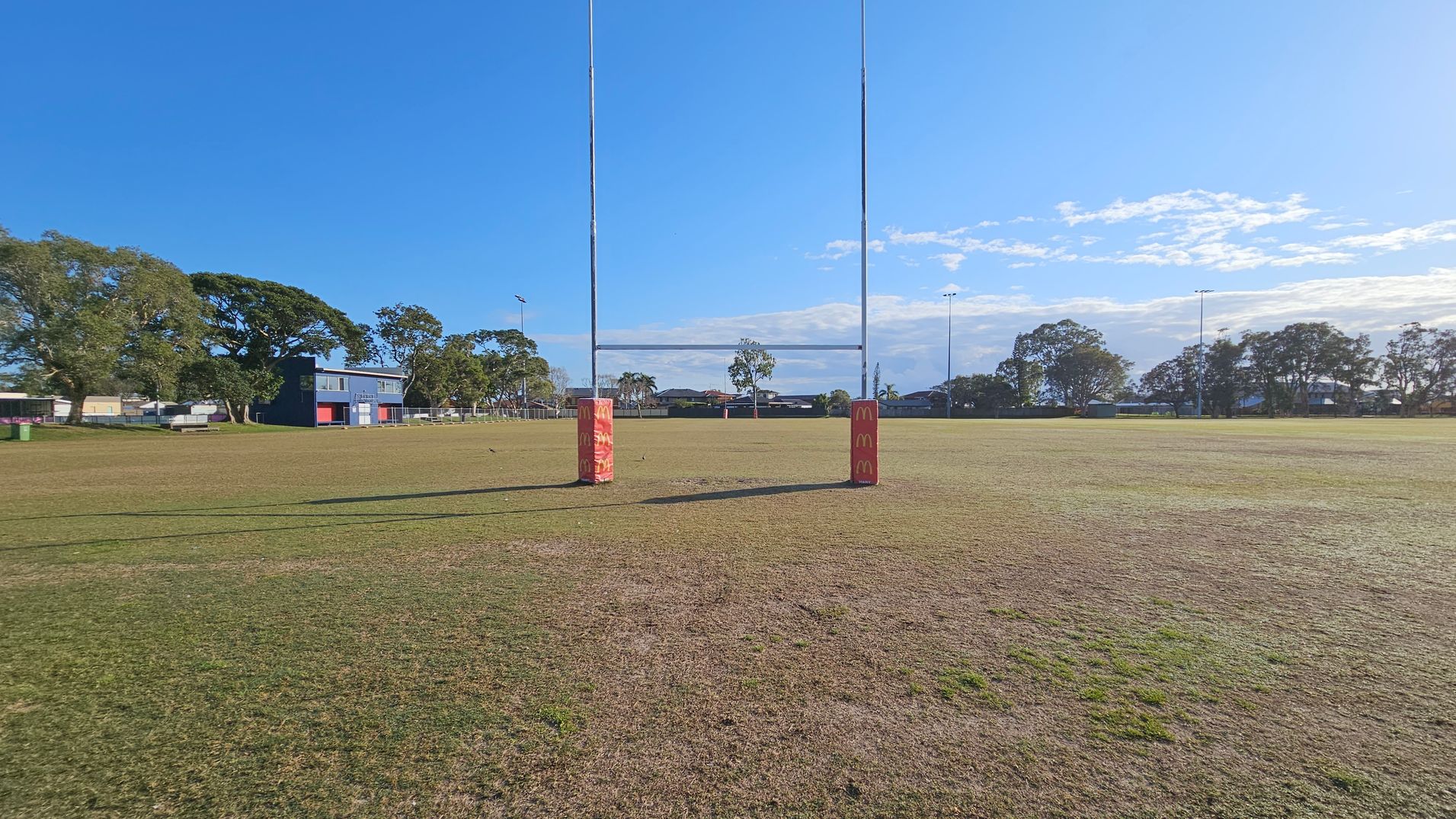 A rugby field with two tall goalposts in the centre, each wrapped with red padding featuring a logo. The grass is patchy and brown in some areas. To the left, there is a blue building with red doors, surrounded by trees. More trees and a few houses are visible in the background. The sky is clear with some clouds on the right.