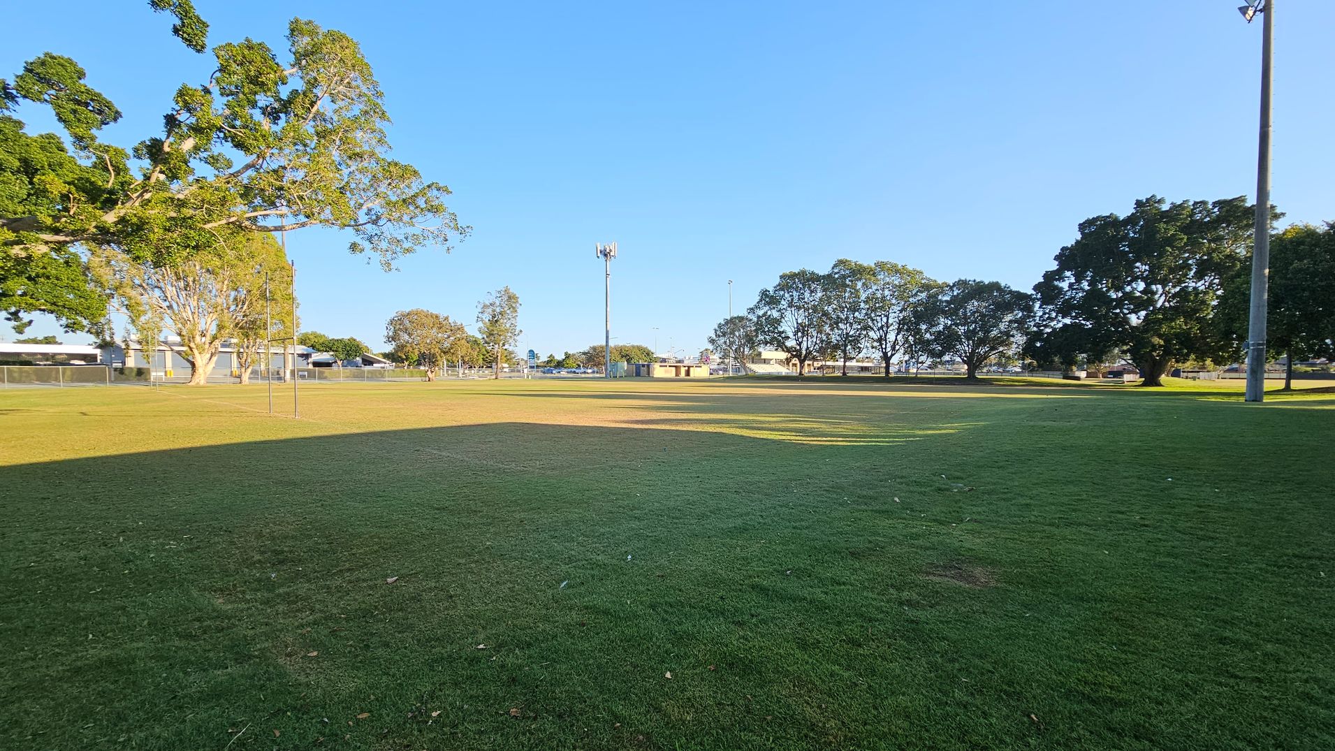 A grassy field with patches of brown and green. On the left, there are large trees with dense foliage and a metal fence in the background. In the centre, a tall light pole and a small building are visible. On the right, more trees and another light pole can be seen. The sky is clear and blue, casting long shadows across the field.