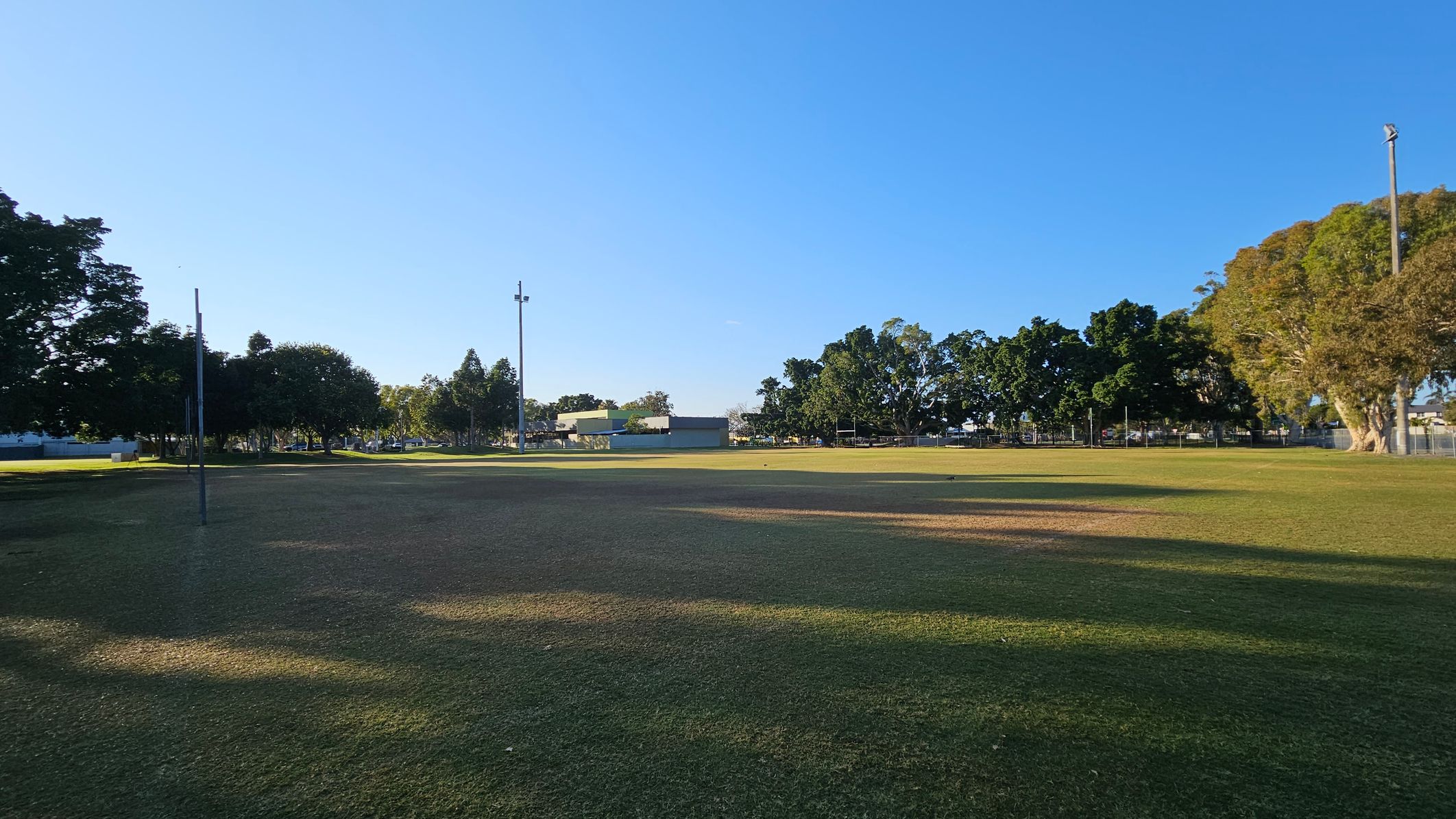 A grassy field with patches of brown and green grass. In the background, there are several tall trees and a few buildings partially visible. Two tall light poles are positioned on the left and right sides of the field. The sky is clear and blue, and the shadows of the trees stretch across the grass.