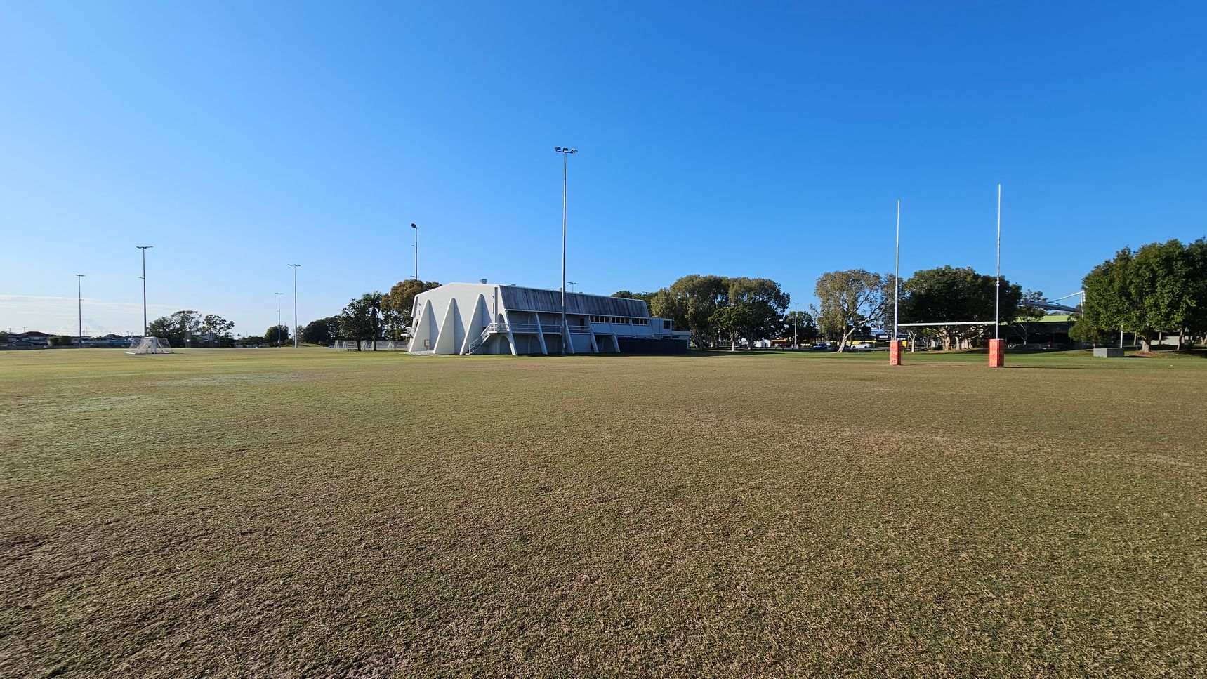 A large, open sports field with a brownish grass surface. On the left, there are several tall floodlights and a few small goalposts. In the centre, a white building with a slanted roof and triangular supports is visible. On the right, a set of rugby posts with red padding stands prominently. Trees and additional structures are in the background, providing a natural border to the field. The sky is clear and blue.