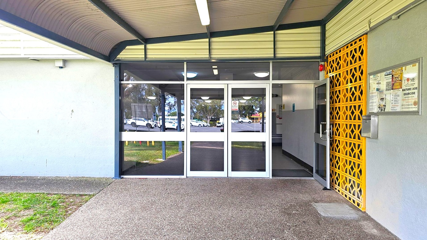 A building entrance with glass double doors in the centre, reflecting parked cars and trees outside. To the right, a yellow lattice panel and a noticeboard with various posters. The left side features a plain grey wall. The floor is a mix of concrete and grass, and the ceiling is covered with a corrugated metal awning. A fluorescent light fixture is mounted on the ceiling.