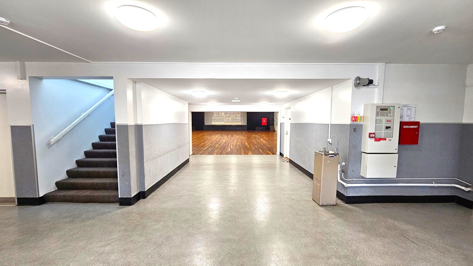 A hallway with a green and grey floor and white ceiling with circular lights. On the left, a staircase with a dark carpet leads upwards. On the right, a wall-mounted fire alarm system and a water fountain are visible. In the centre, the hallway opens into a large room with a wooden floor and a stage at the far end. The walls are grey with a white upper section.