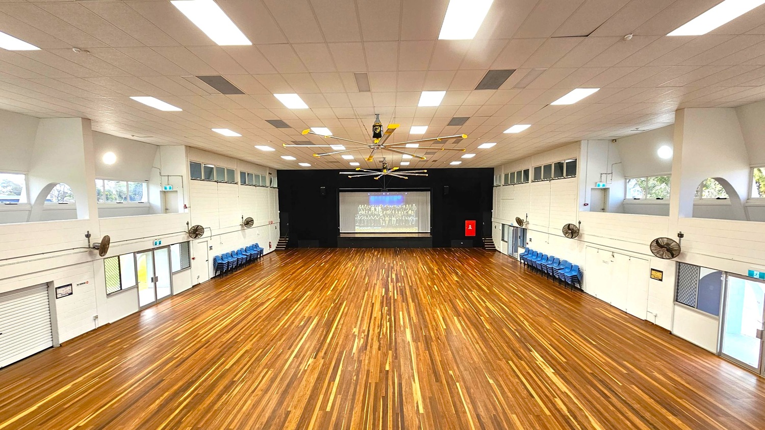 A large hall with a polished wooden floor and a high ceiling with bright lights. In the centre, a stage with a screen displaying an image. On both sides, rows of blue chairs are stacked against the walls. The walls are light-coloured with several fans and windows. The ceiling features a large fan. Doors and emergency exits are visible on the sides.