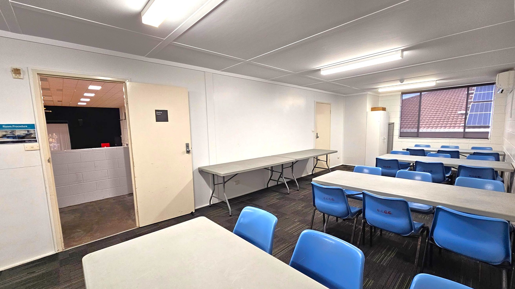 A room with white walls and a grey ceiling featuring fluorescent lights. On the left, an open door leads to another room with a purple half-wall. A long folding table is against the left wall. On the right, several tables are arranged with blue plastic chairs around them. A large window shows a view of a tiled roof with solar panels. The floor is covered with dark carpet tiles.