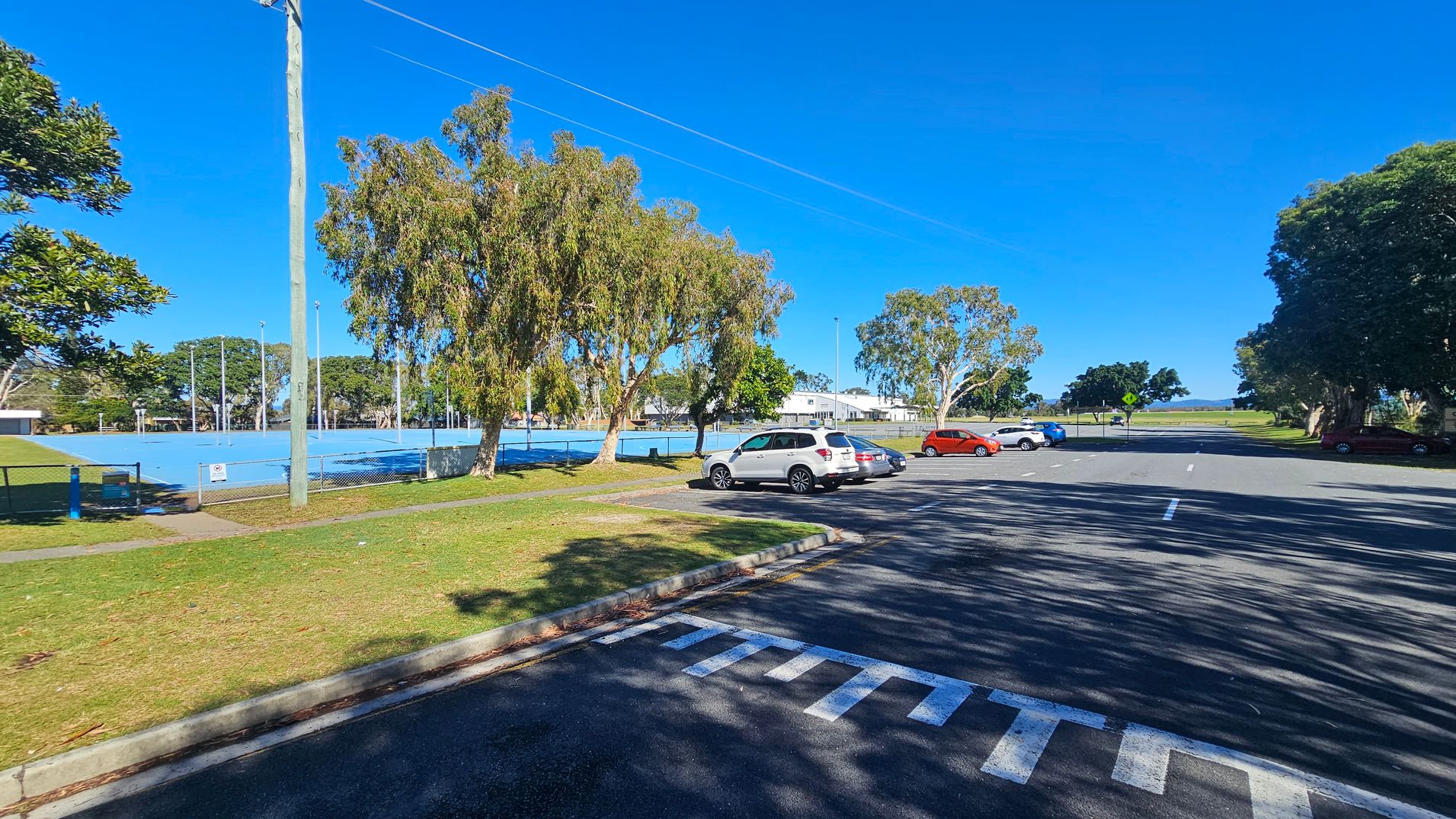 A parking area with several cars is on the right, with a few trees providing shade. On the left, there is a fenced sports court with a blue surface, surrounded by more trees and a grassy area. A tall utility pole stands near the court. In the background, a white building is visible, and beyond that, open fields stretch towards the horizon. The ground is paved, and the sky is clear.