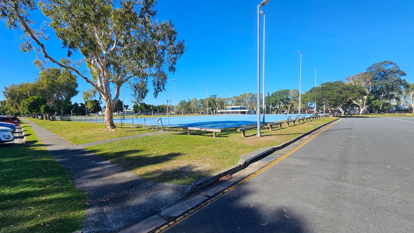A paved road runs along the right side, bordered by a yellow line. On the left, a row of parked cars is adjacent to a grassy area with a footpath and several large trees. In the centre, a fenced sports court with tall floodlights is visible. Benches are placed along the perimeter of the court. In the background, more trees and a building can be seen. The sky is clear and blue.