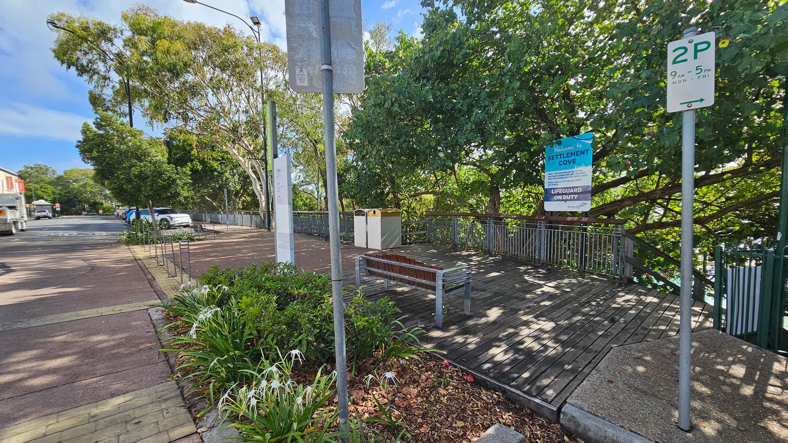 A street scene with a wooden deck area on the right, featuring a bench and two bins. A sign reads "Welcome to Settlement Cove" with lifeguard information. Trees with dense green foliage provide shade. On the left, a paved footpath runs alongside the road, with parked cars and a truck visible. The area is landscaped with plants and flowers. Parking signs are visible, and the sky is clear.