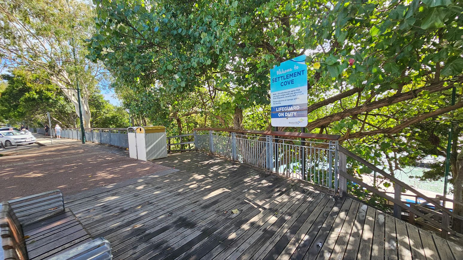 A wooden boardwalk with metal railings runs along the right side, partially shaded by dense green foliage. A sign reading "Welcome to Settlement Cove" is visible among the trees. On the left, a paved path with parked cars and a few people walking. A bin with a yellow lid is near the railing. The background features more trees, and glimpses of water can be seen through the foliage. Benches are positioned along the boardwalk.