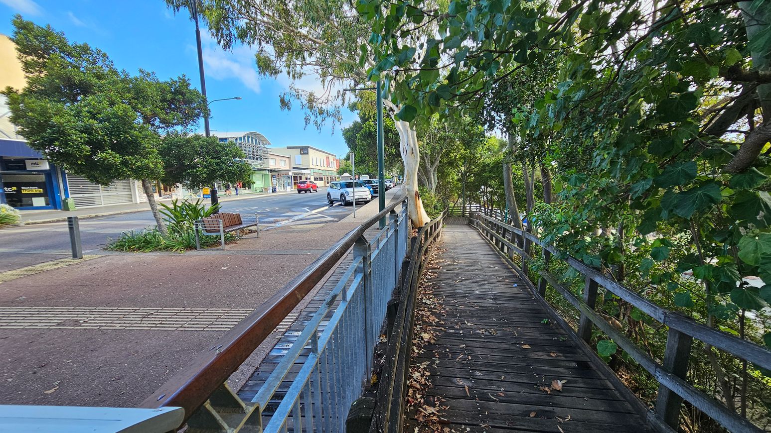 A wooden walkway on the right is bordered by lush green trees and a metal railing. On the left, a street with parked cars and a row of shops, including a blue storefront, is visible. A bench and a small garden with a tree are situated near the pavement. The ground is a mix of concrete and reddish paving, and a tall streetlamp is present. The scene is urban with a touch of greenery.
