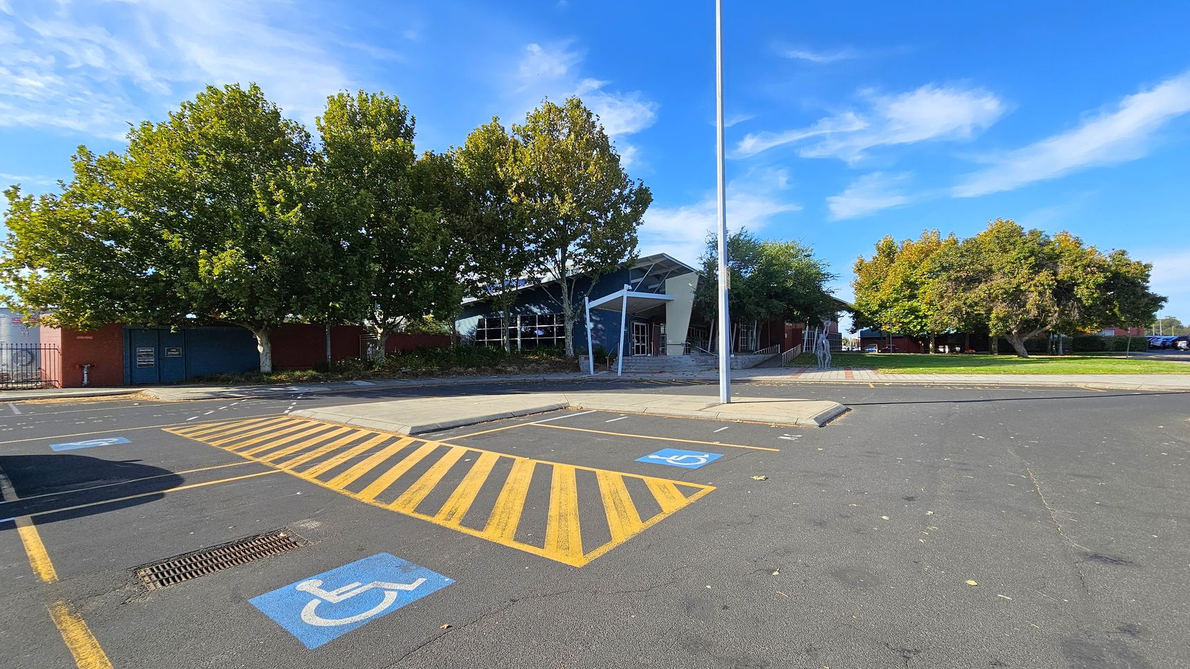 A parking area with marked disabled parking spaces in the foreground, featuring blue symbols and yellow striped lines. In the middle, a modern building with a slanted roof and large windows is partially obscured by trees. The background has more trees and a grassy area. A tall streetlight stands near the centre. The pavement is dark grey, and the sky is clear with some clouds.