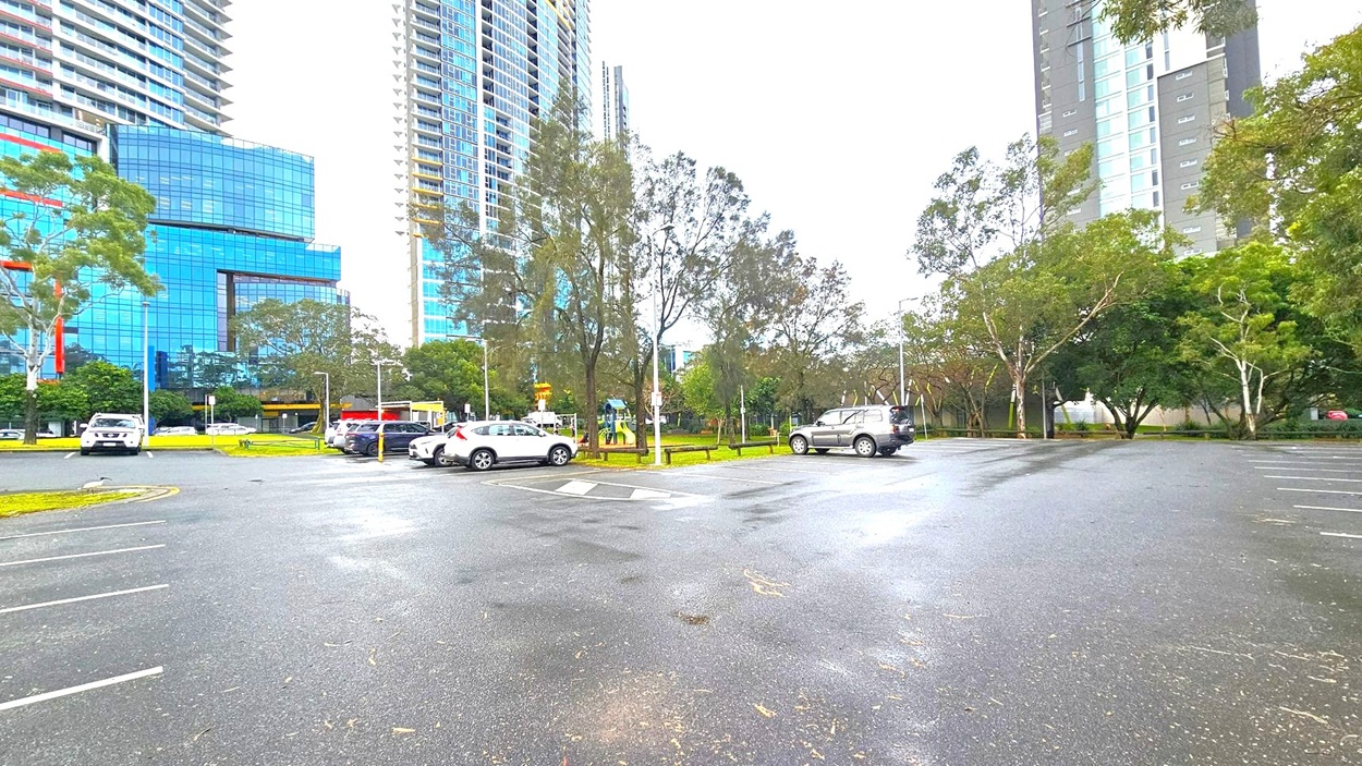 A car park with several vehicles is in the foreground, with marked parking spaces on the asphalt. In the background, there are tall buildings with reflective glass windows on the left and right. Trees with sparse foliage are scattered throughout the area, and a small playground is visible among the trees. The scene is set in an urban environment with a mix of natural and architectural elements.