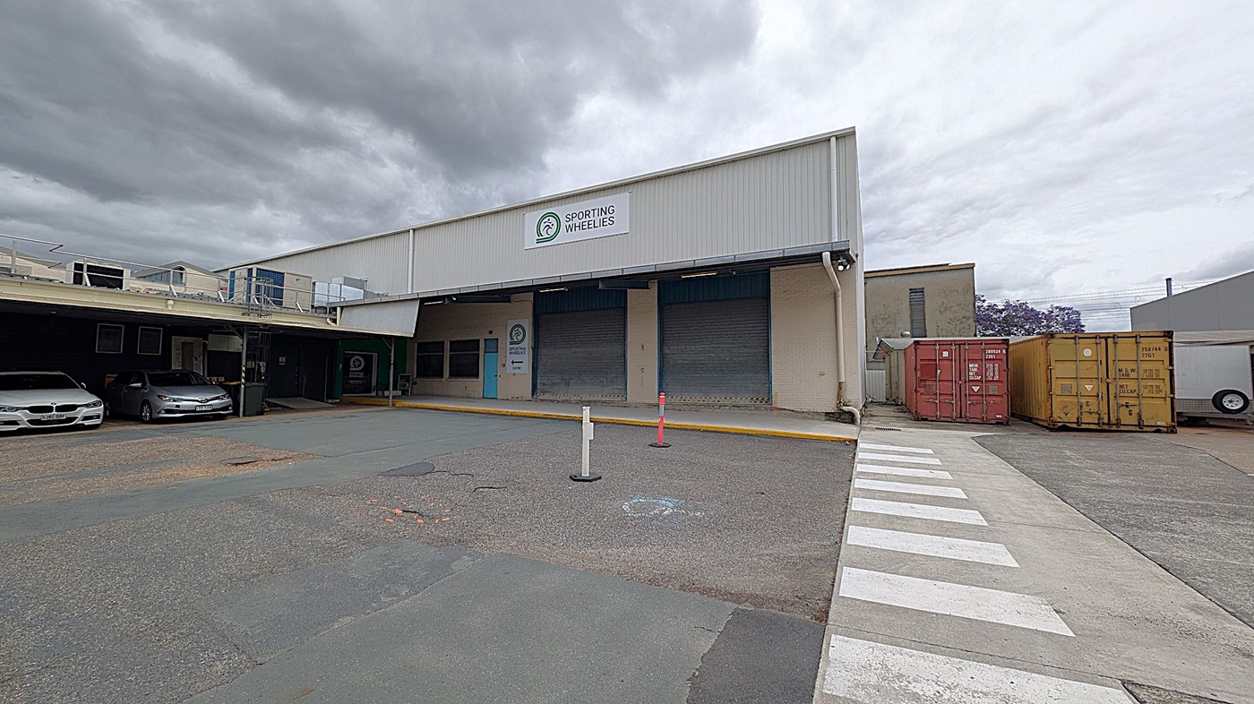 A warehouse with a sign reading "Sporting Wheelies" is in the centre. On the left, two cars are parked under a covered area. On the right, there are two shipping containers, one red and one yellow. A zebra crossing leads to the entrance, and there are two bollards in front of the building. The ground is paved, and the building has a corrugated metal exterior.