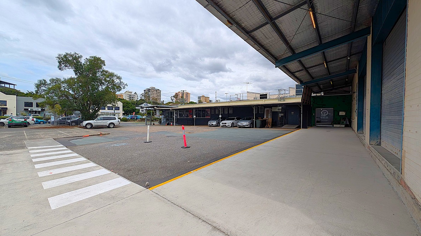 A covered loading area is on the right with a concrete floor and a metal roof. Several cars are parked in front of a low building in the middle distance. On the left, a large tree stands near a row of parked cars. A zebra crossing leads from the foreground towards the tree. In the background, there are taller buildings visible beyond the low structure.