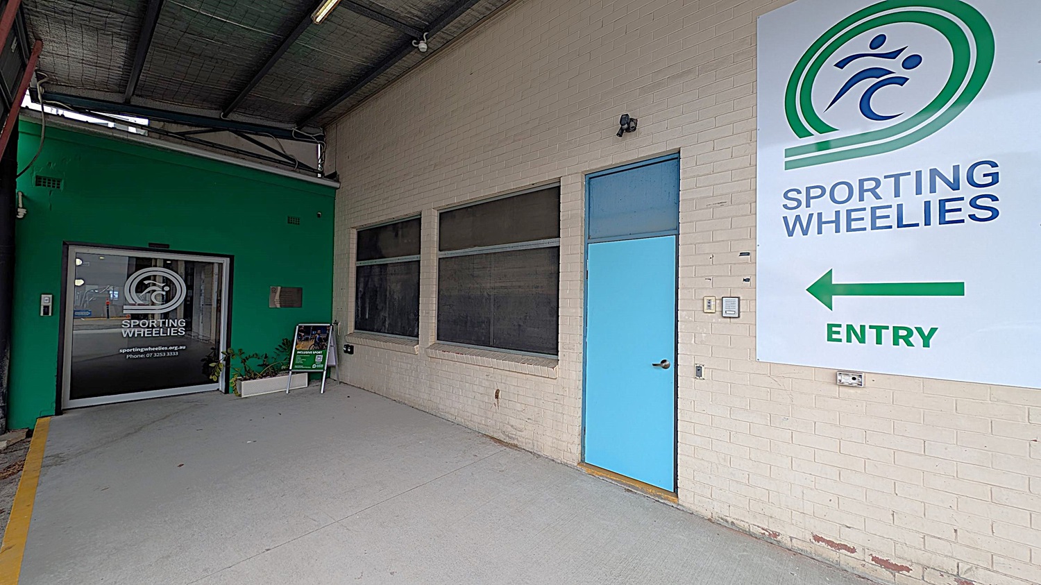 A covered entrance area with a concrete floor. On the right, a large sign on a light brick wall reads "Sporting Wheelies" with an arrow pointing left. Below the sign is a blue door. On the left, a green wall features a glass door with the Sporting Wheelies logo and contact information. A small planter and a signboard are near the entrance. The ceiling is industrial with exposed beams and lighting.