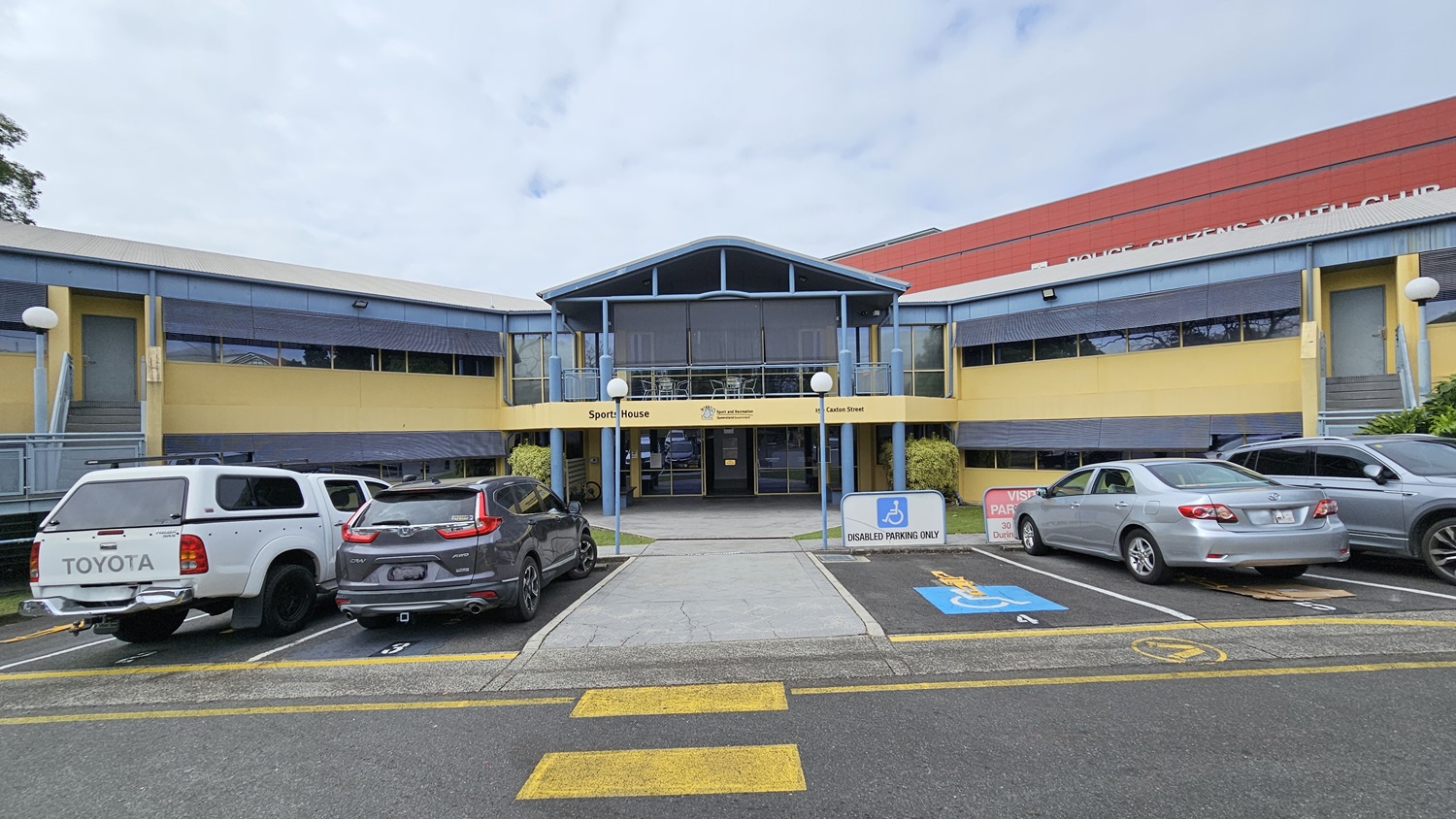 A paved driveway curves from the foreground to the background, flanked by yellow lines and a pedestrian crossing. On the left, there are parked cars and tall grass. In the middle, a two-storey building with a blue and yellow facade, featuring large windows and a staircase. On the right, a parking area with more cars, a sign for authorised parking, and a yellow parking barrier. Trees and bushes surround the area, providing greenery.