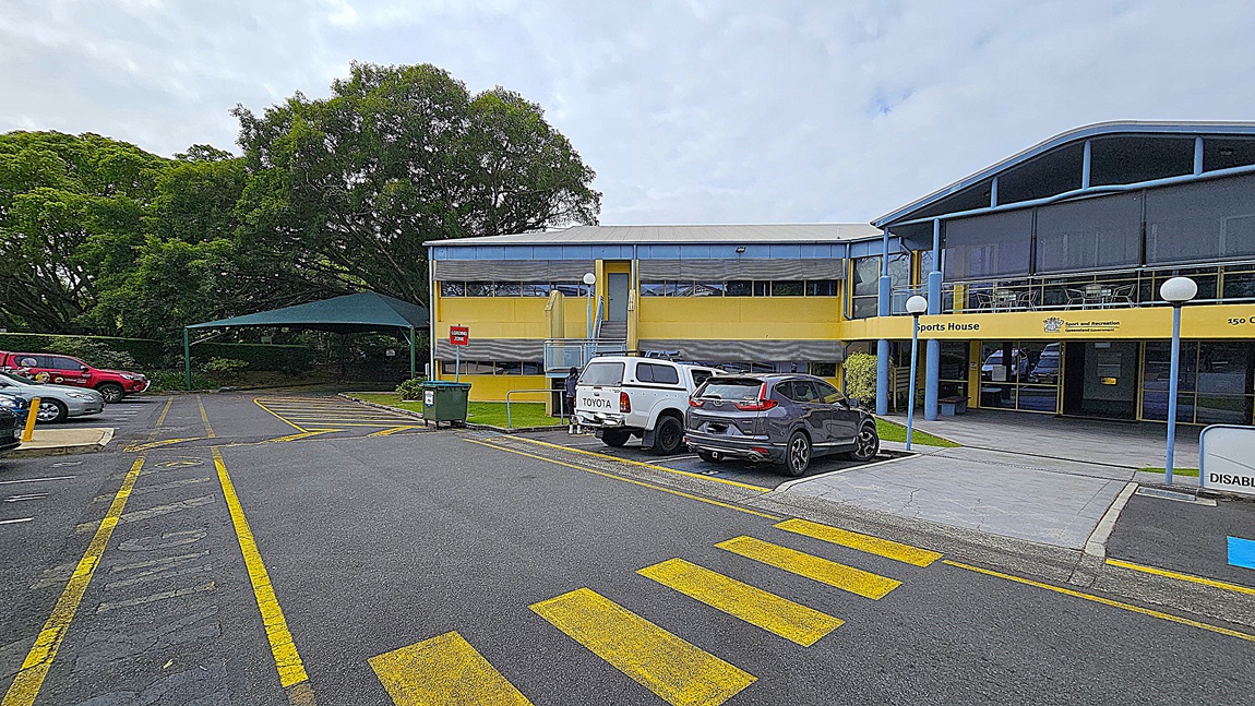 A two-storey building with a blue and yellow facade is in the centre, labelled "Sports House." The foreground has a paved area with yellow pedestrian crossing lines. On the left, there are parked cars, including a white Toyota, and a green bin near a covered area with trees. On the right, more parked cars, including a grey SUV, and a sign for disabled parking. The background includes dense green trees. The floor is asphalt with marked parking spaces and pedestrian lines.