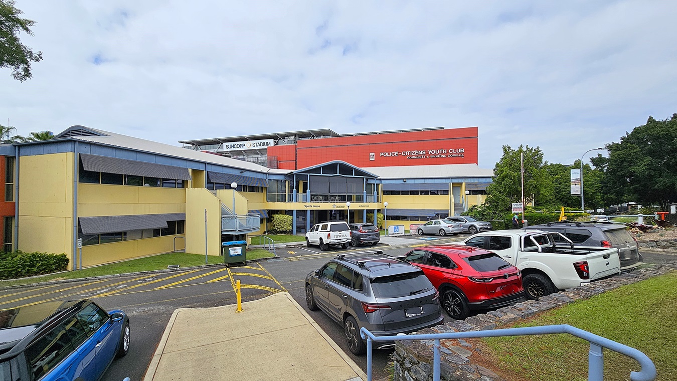 A two-storey building with a blue and yellow facade is in the centre, labelled "Police-Citizens Youth Club." The foreground has a paved area with yellow pedestrian crossing lines and a concrete walkway. On the left, a blue car is parked near a green hedge. On the right, several parked cars, including a grey SUV, a red car, and a white pickup truck, are adjacent to a grassy area with a stone border. The background features a red building with "Suncorp Stadium" signage.