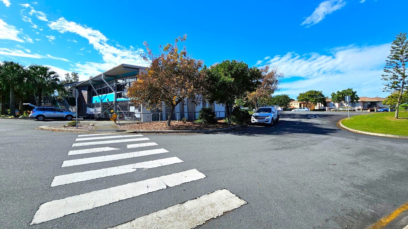 A zebra crossing leads to a building with a modern design on the left, featuring large windows and a slanted roof. Several trees with autumn foliage are in front of the building. A silver car is parked nearby. On the right, a white vehicle is parked along a curved road, with a grassy area and more trees. In the background, there are residential houses with tiled roofs. The sky is clear with a few clouds.