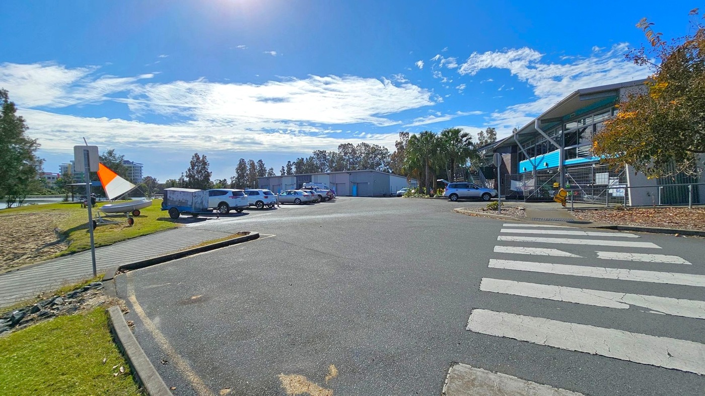 A car park with several vehicles is in the centre, surrounded by trees and a building on the right with a blue facade. A pedestrian crossing is visible in the foreground. On the left, a grassy area with a small boat and a trailer is near a path. A sign with an orange and white flag is also on the left. The background features more trees and a few buildings.