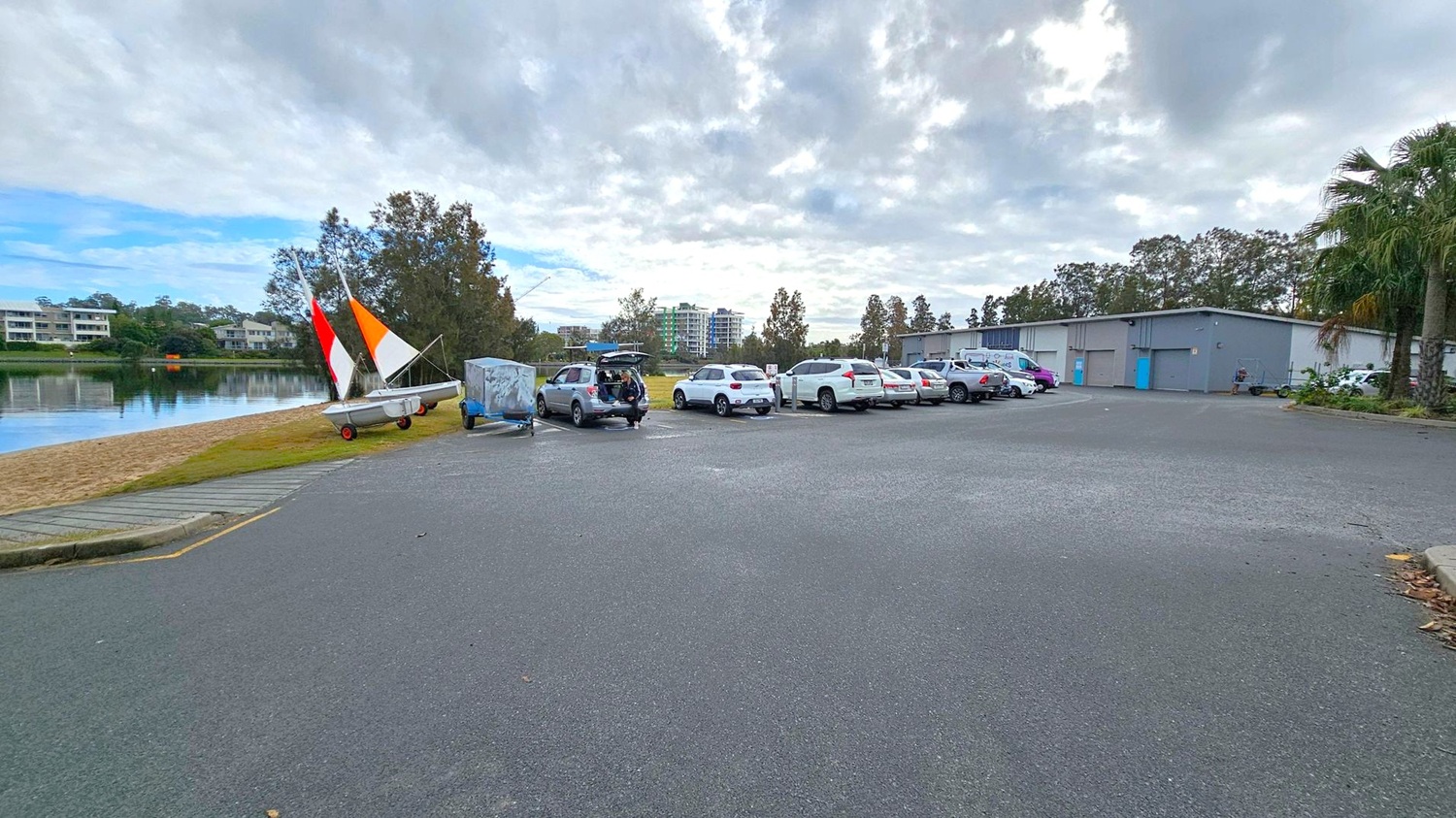 A car park with several vehicles is on the right, with a row of white and silver cars. In the middle, a car has its boot open. On the left, a small sailboat with red, orange, and white sails is on a grassy area near a sandy shore. In the background, there are trees and a few multi-storey buildings. A large grey building with blue doors is on the right. The ground is paved, and there are some palm trees on the far right.