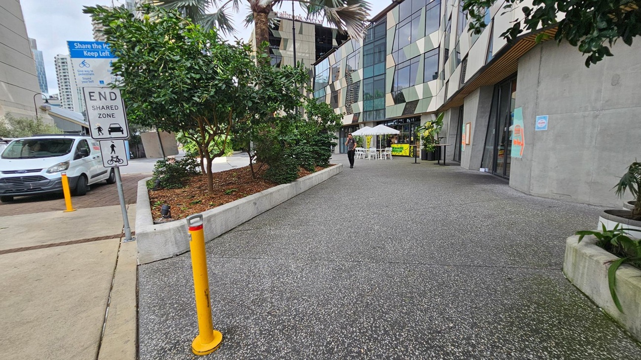 A city street scene with a modern building on the right featuring geometric glass panels. In front of the building, there are outdoor tables with white umbrellas. A person walks along the pavement. On the left, a white van is parked near a sign indicating the end of a shared zone. The foreground has a textured grey pavement with yellow bollards, and there are green plants in raised beds along the path.