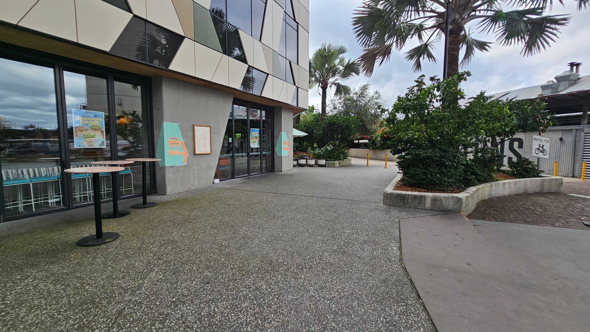 A modern building with geometric patterns on the facade is on the left, featuring large glass doors and posters. Two round tables stand outside. The ground is a mix of green and grey textured surfaces. On the right, there are landscaped areas with bushes and a palm tree. A sign warns to watch for bicycles. In the background, there is a metal structure with a partially visible word. The sky is overcast.