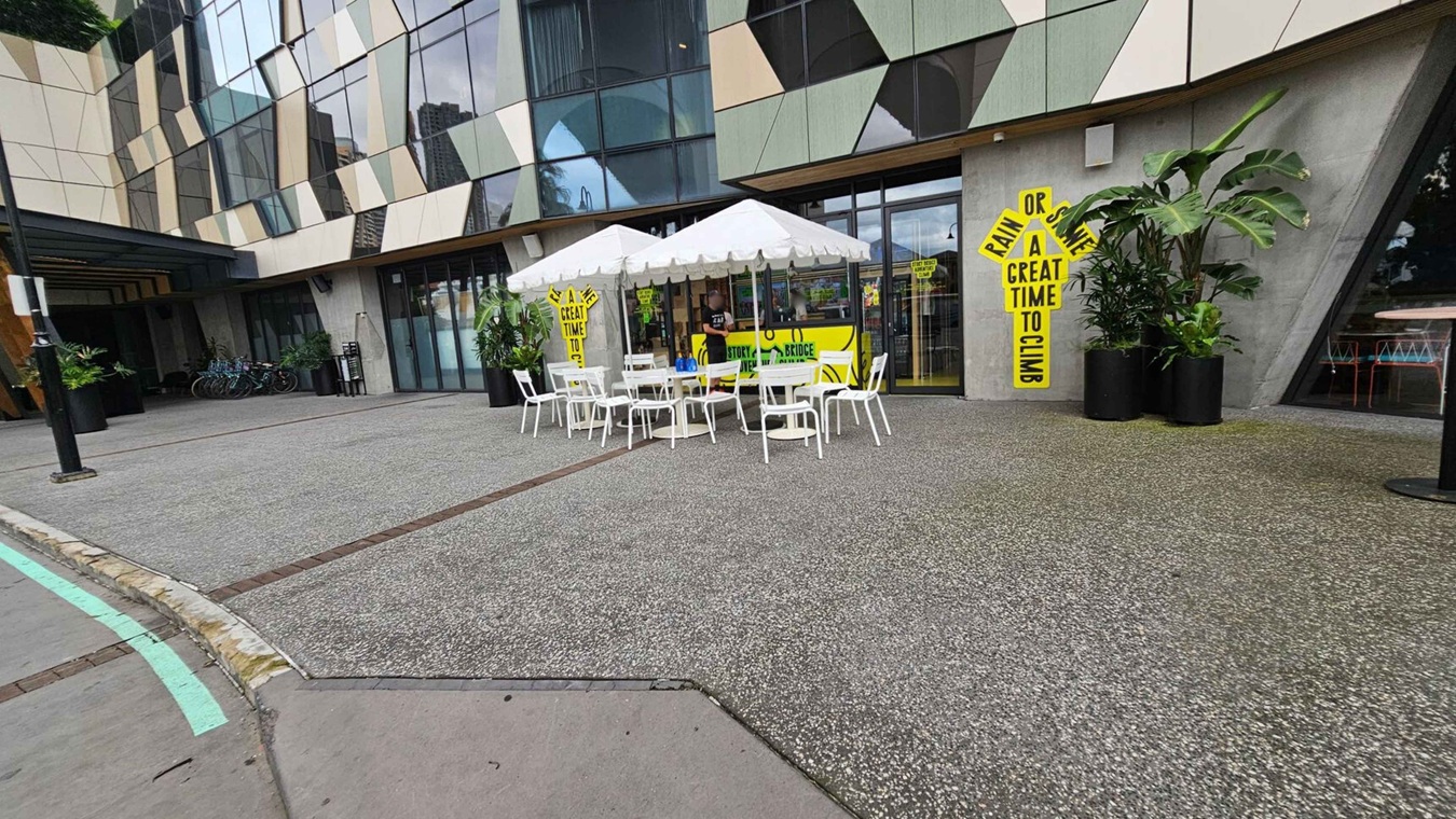 A modern building with a geometric facade featuring large glass windows and patterned panels. In front, there is a seating area with white tables and chairs under white umbrellas. Bright yellow signs with black text are displayed near the entrance. Several potted plants are placed around the area. Bicycles are parked to the left, and the ground is a mix of grey and green textured surfaces.