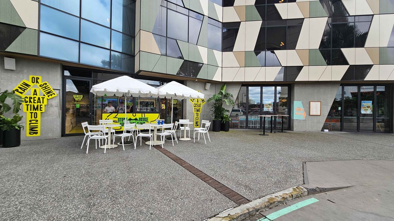 The foreground features a patio area with white tables and chairs under white umbrellas. On the left, a bright yellow sign reads "Rain or Shine A Great Time to Climb." The building facade has a geometric pattern of glass and panels in various shades. Large windows and doors are visible, with posters and signs inside. Potted plants are placed near the entrance. The ground is a mix of concrete and green artificial turf.