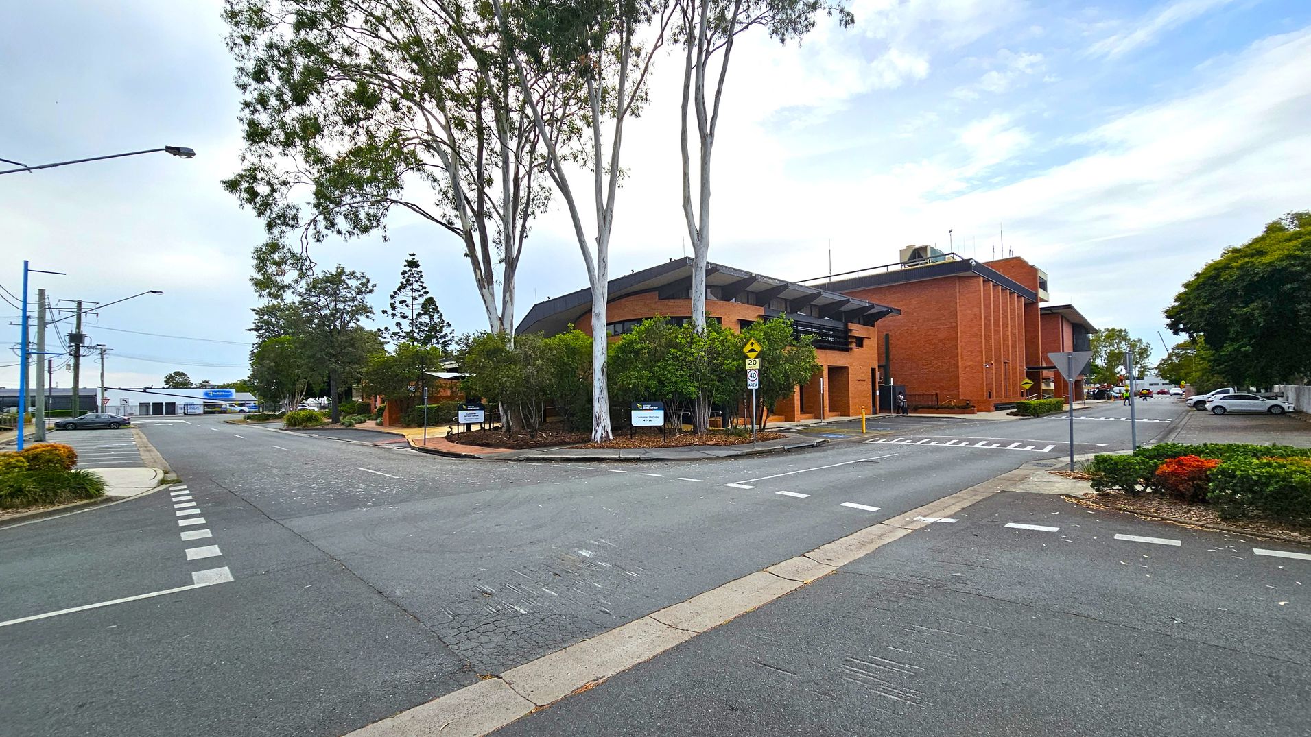 A street intersection with a brick building on the right, featuring a modern design and surrounded by trees. The road has marked pedestrian crossings and a give-way sign. On the left, there are more trees and a few parked cars. The background shows additional buildings and a clear sky. The foreground has a paved road with visible lane markings.