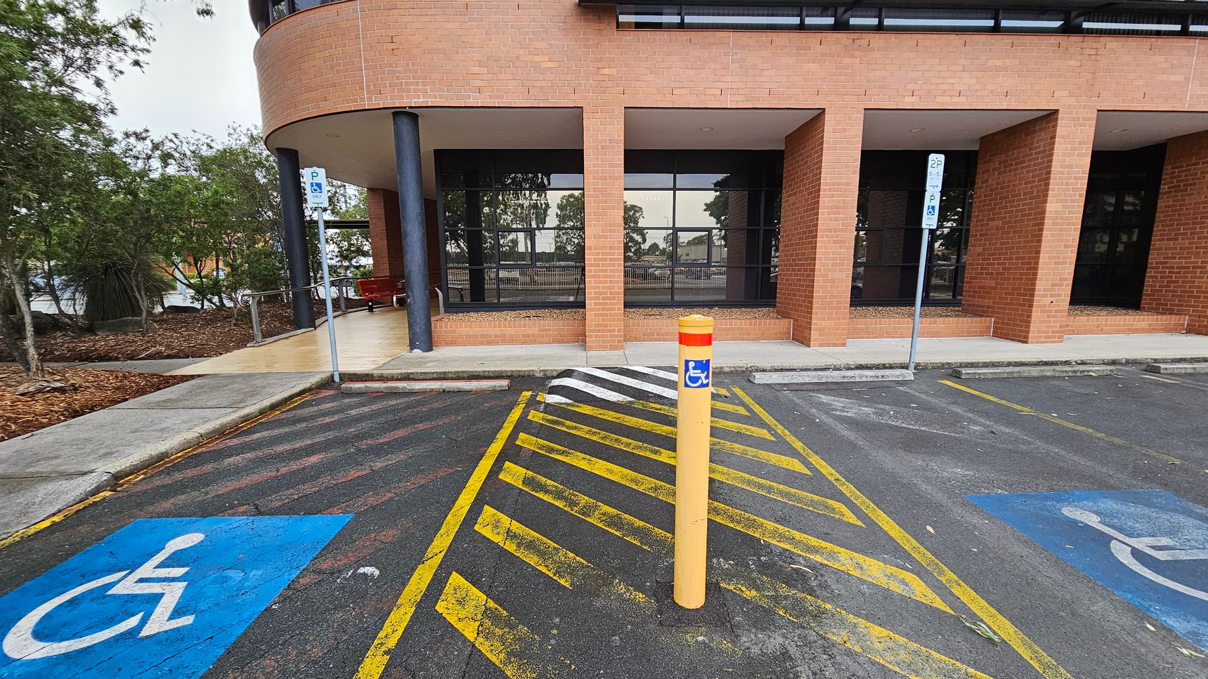 A parking area with two marked disabled parking spaces on either side, featuring blue wheelchair symbols. In the centre, a yellow bollard with a blue disabled sign stands on a yellow-striped zone. The background shows a brick building with large windows and a covered walkway supported by black columns. To the left, there are trees and a landscaped area with mulch. The ground is asphalt, and the ceiling of the walkway is light-coloured.