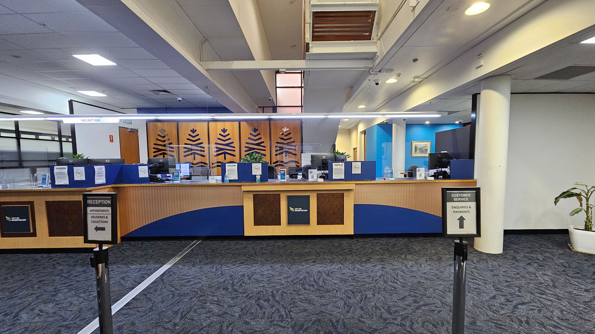 Reception area with a long wooden counter featuring blue accents and decorative panels. Behind the counter are computer monitors and signs. Two signs in the foreground direct visitors to reception and customer service. The floor is carpeted in a dark pattern, and the ceiling has recessed lighting. A potted plant is visible on the right, and the walls are painted in blue and white tones. An exit sign is above the counter.