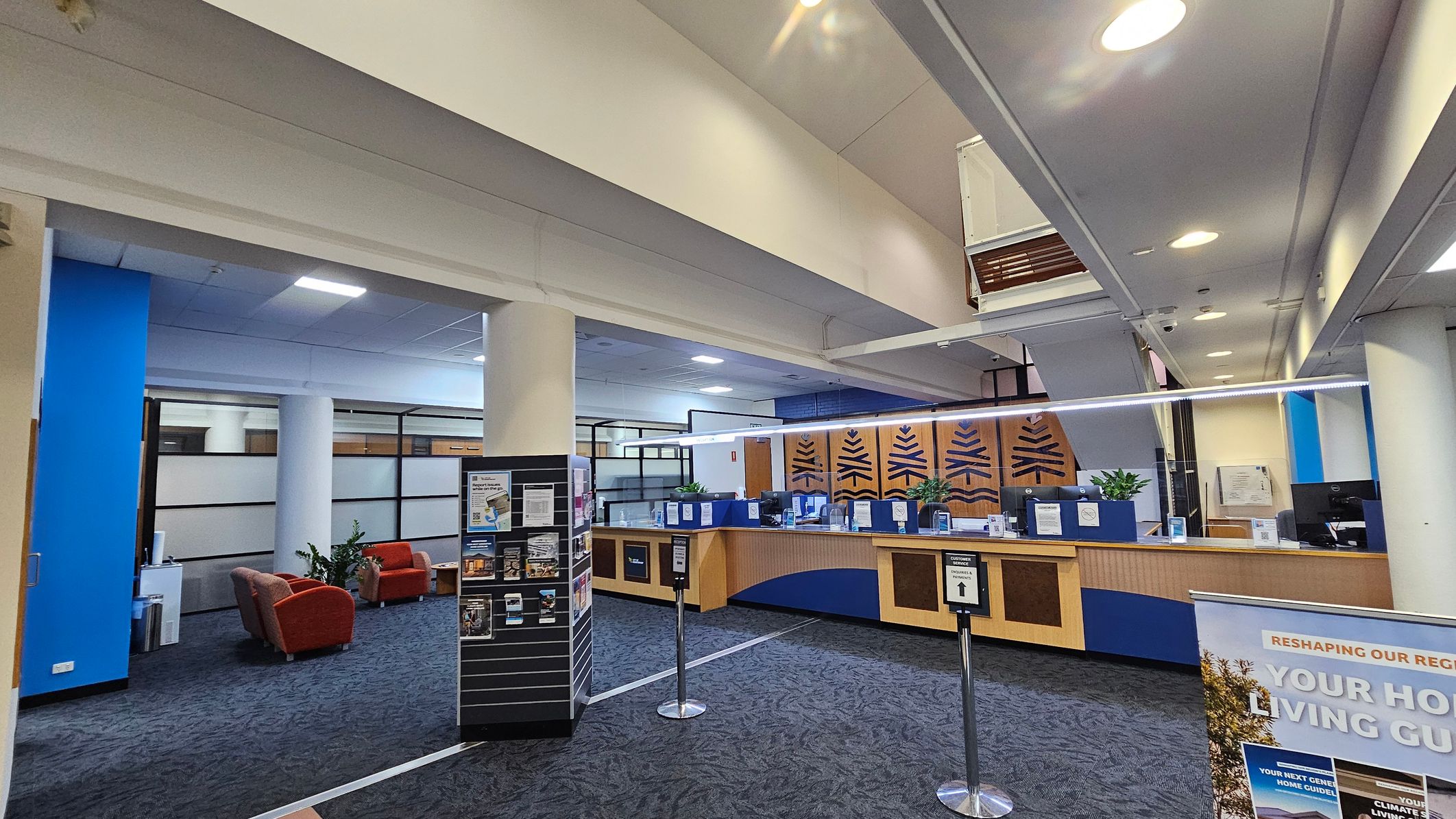 A spacious interior with a reception area on the right, featuring a wooden counter with blue accents and decorative panels. Potted plants are placed on the counter. In the foreground, a sign and a brochure stand are visible. On the left, there are red armchairs and a small table near a blue wall. The floor is carpeted in a dark pattern, and the ceiling has recessed lighting. Large windows with frosted glass panels are in the background.
