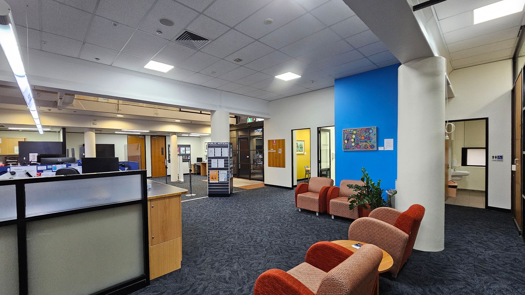 A spacious office area with a blue accent wall on the right, featuring a colourful artwork. In front of the wall are several cushioned chairs and a small table. A plant is placed nearby. On the left, there are workstations with computers and a partitioned desk. The floor is carpeted in a dark pattern, and the ceiling has recessed lighting. A public notice board stands near the centre, and there are doors leading to other rooms.