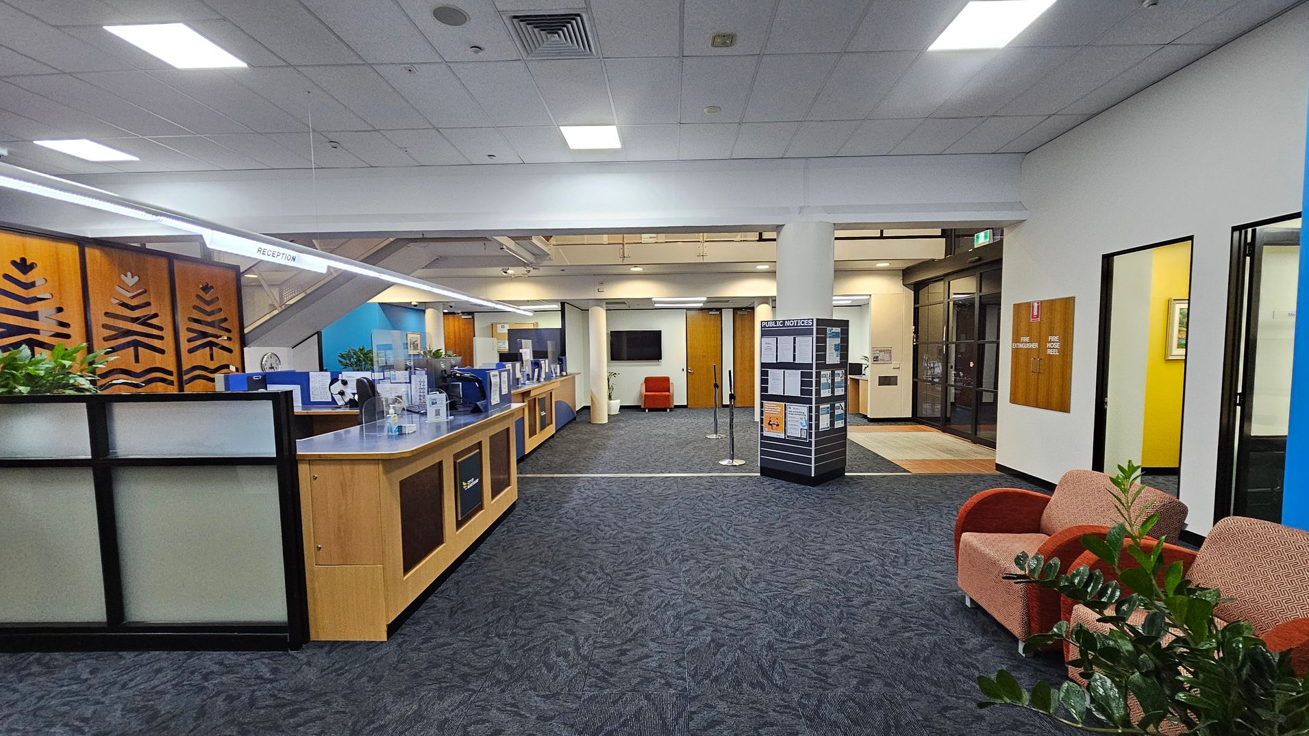 Reception area with a wooden counter on the left, featuring decorative panels and potted plants. A sign above reads "RECEPTION." In the centre, a display board with public notices stands on a carpeted floor. To the right, two upholstered chairs are placed near a plant. The background includes a wall-mounted TV and wooden doors. The ceiling has recessed lighting, and the floor is covered with patterned carpet tiles.