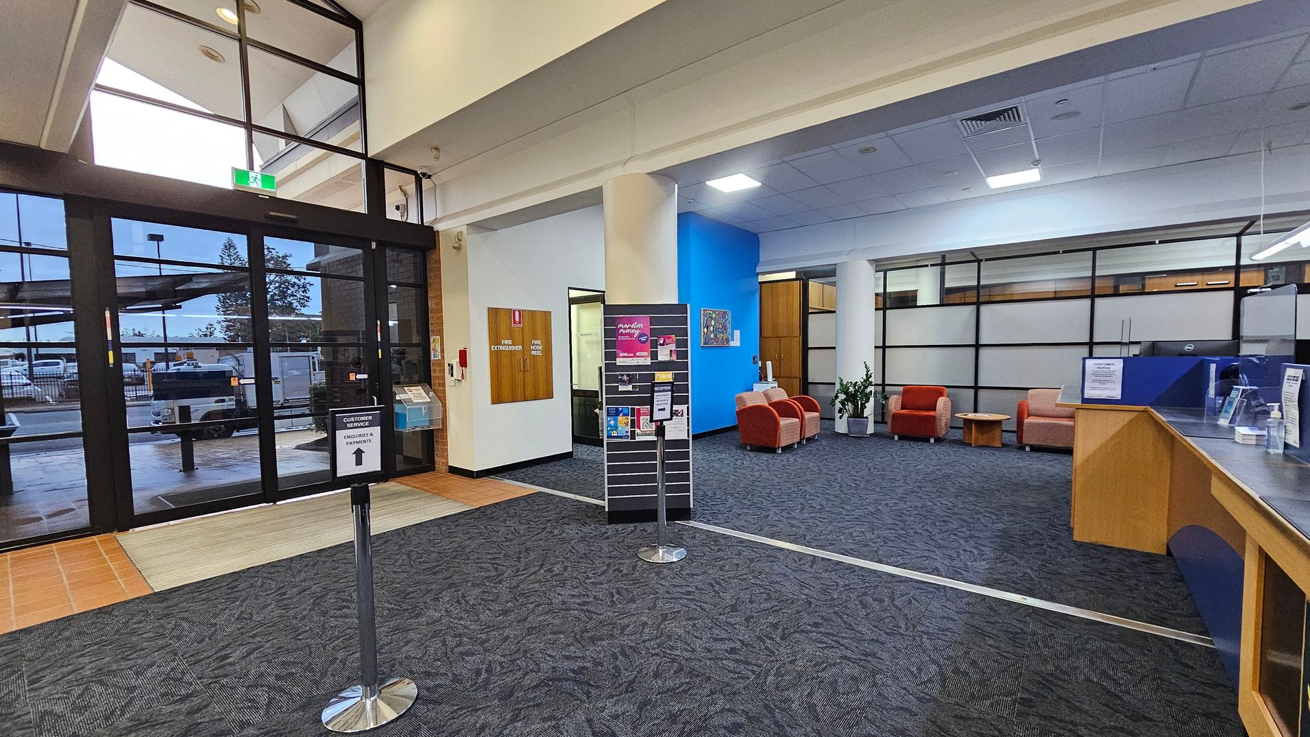 Entrance area of a public building with large glass doors on the left, leading outside. A sign directs to customer service. In the centre, a display stand holds brochures. To the right, a waiting area with red and beige chairs, a small table, and potted plants. The floor is carpeted in dark grey, and the ceiling is fitted with lights and air vents. A wooden counter with a clear screen is on the far right.