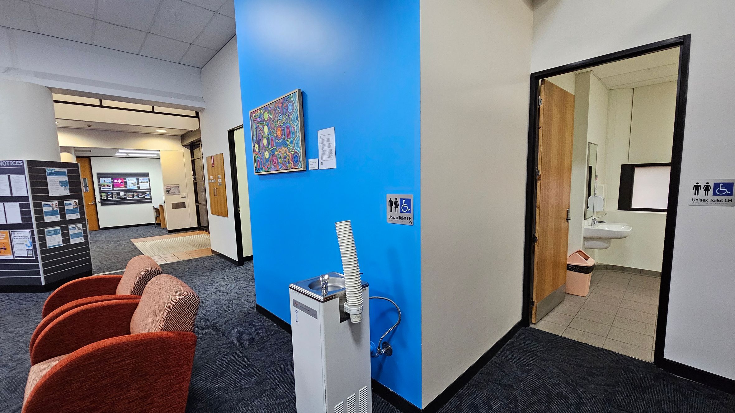 A hallway with a blue accent wall in the centre, featuring a colourful artwork. Below the artwork is a water fountain with stacked cups. To the right, an open door leads to a unisex accessible toilet, visible with a sink and bin. On the left, two red chairs are placed on a dark carpeted floor. Further left, a notice board displays various papers. In the background, a corridor with a light-coloured floor and a wooden door is visible. The ceiling is tiled.