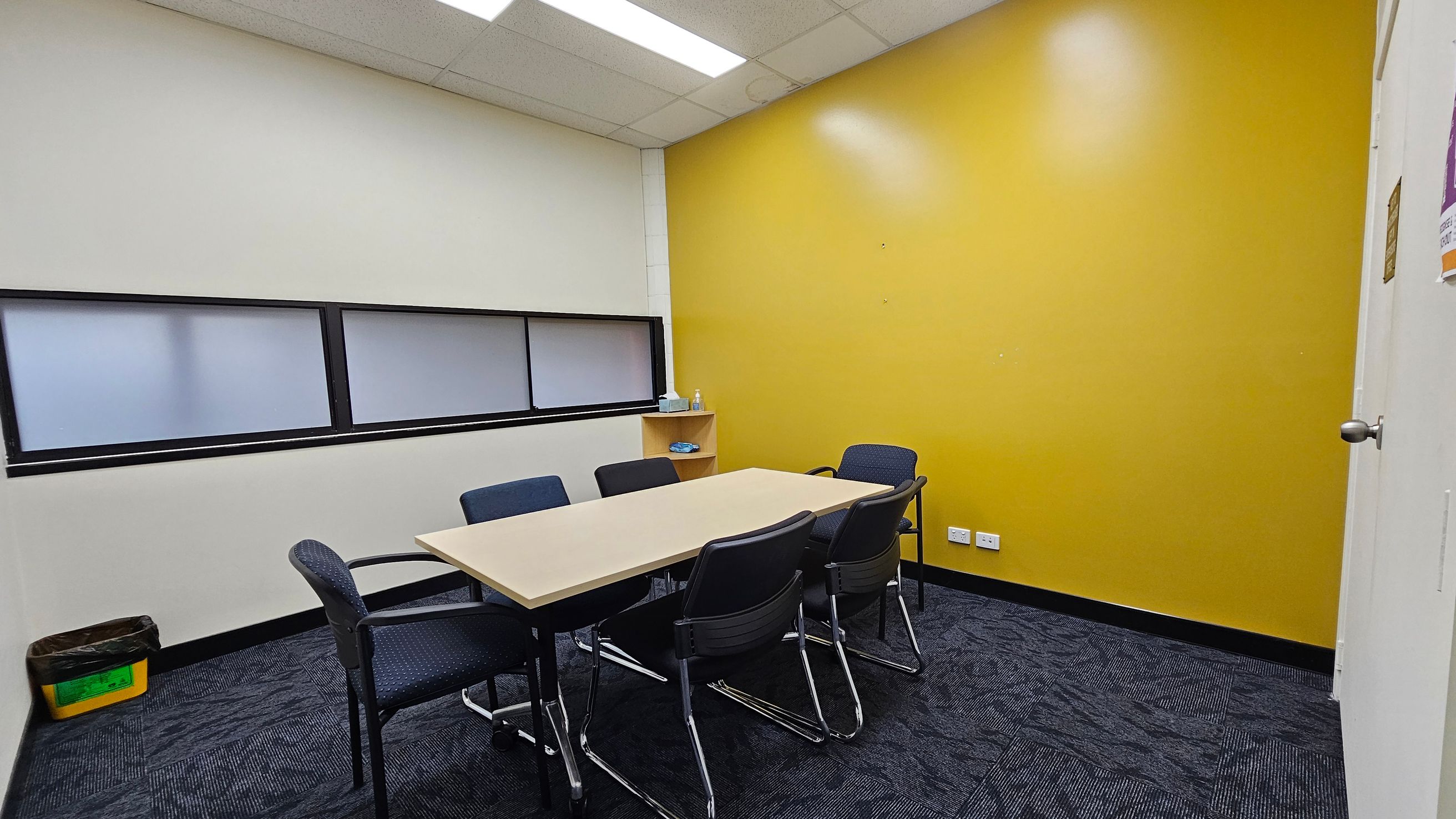 A small meeting room with a rectangular table surrounded by six chairs. The left wall is white with a horizontal frosted window, and a yellow bin is on the floor. The right wall is bright yellow with a small shelf holding a bottle and some items. The floor is covered with dark patterned carpet tiles. The ceiling has white panels with fluorescent lights.