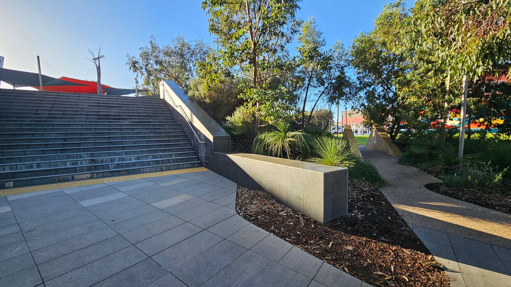 Wide stone steps on the left lead upwards, bordered by a low wall. A path on the right curves through a landscaped area with trees and shrubs. The ground is paved with large tiles, and there is a textured yellow strip at the base of the steps. In the background, colourful structures and a clear blue sky are visible.