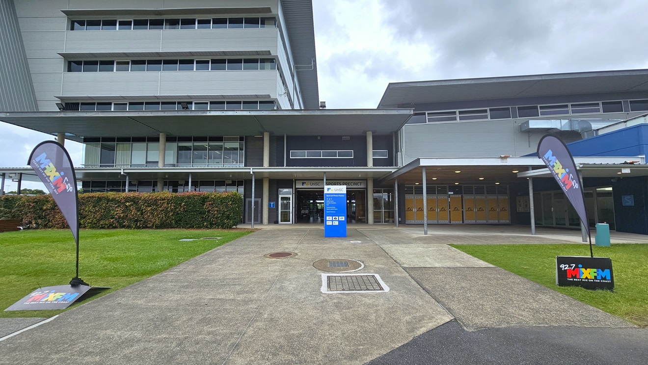 A multi-storey building is bordered at the base by a hedge. Centrally, a concrete pathway leads to an entrance marked by a blue sign and a metal grate on the ground. To the right, the building extends with large windows and a covered walkway. The upper floors, visible in the background, are clad in glass and metal panels. The foreground features concrete pavement and a grassy area, with feather banners and signage visible. Trimmed shrubs line the front to the left.