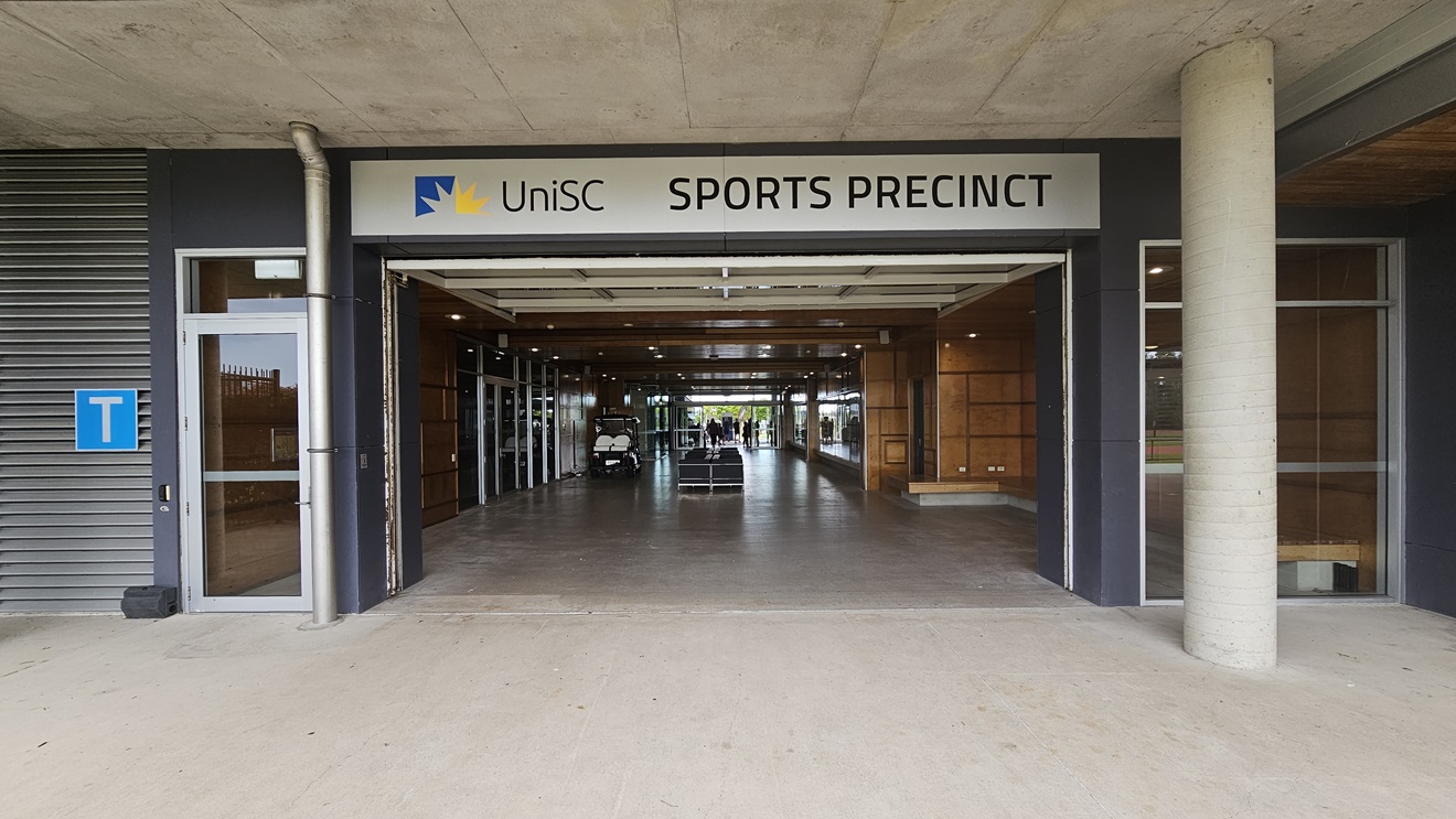 On the left, there is a grey wall with a blue "T" sign and a glass door. In the middle, an open entrance leads into a hallway with wooden panels and glass doors, with a sign above reading "UniSC Sports Precinct." The right side features a concrete pillar and a section of the building with dark walls. The background includes the interior of the sports precinct with visible lights and a small vehicle. The floor is concrete, and the ceiling is made of exposed concrete panels.