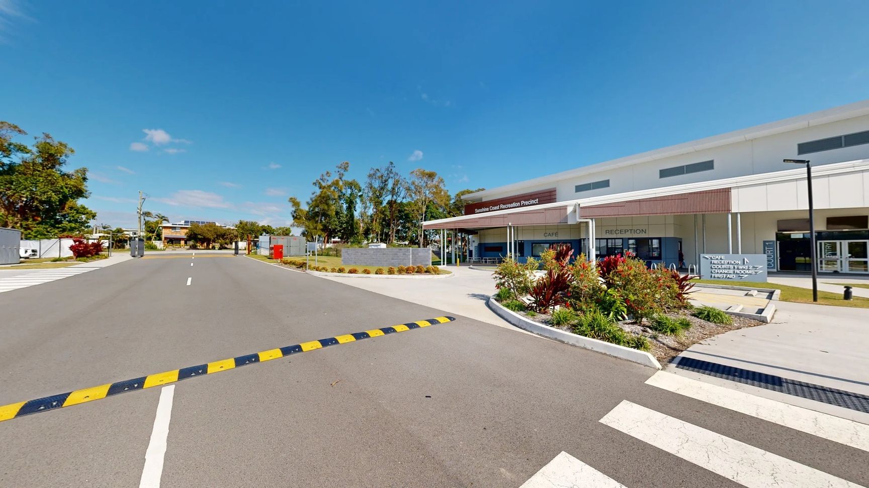 A wide grey asphalt road extends towards a building in the centre. On the left, a landscaped area features various plants and a path leading to an entrance. A red postal box stands prominently. To the right, the modern structure has large glass windows, with visible signage for a café and reception. Brightly coloured foliage outlines the entrance, and a smooth pedestrian crossing indicates safe passage.
