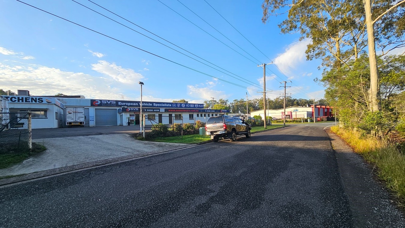 A street scene with a paved footpath in the foreground and a road running parallel. Across the road, there is a row of commercial buildings with signs for "Suncoast Kitchens" and "European Vehicle Specialists." Several cars are parked in front of the buildings. Power lines and poles are visible, casting shadows on the road. The background features trees and a clear sky.