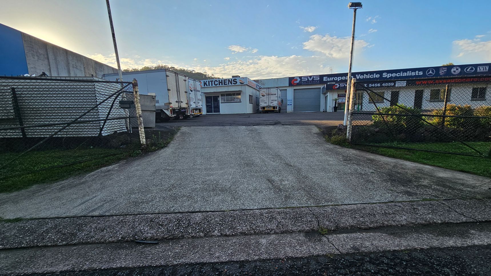 A driveway leads to a commercial area with buildings in the background. On the left, there are two large white trucks parked near a building labelled "KITCHENS." On the right, a building with signage for "SVS European Vehicle Specialists" is visible, along with a closed garage door. A chain-link fence runs along both sides of the driveway, and a tall streetlight stands on the right side. The ground is paved, and there is some grass visible near the fence.