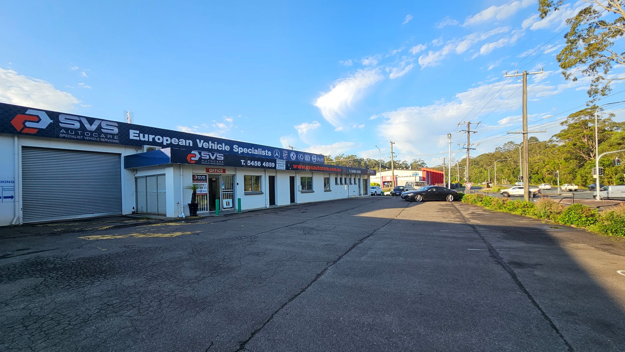 A vehicle service centre with a large sign reading "SVS Autocare European Vehicle Specialists" is on the left. The building is white with a roller door and an office entrance. Several logos of car brands are displayed. On the right, a paved area leads to a road with cars and traffic lights. Trees and greenery are visible in the background, along with power lines and a clear sky.