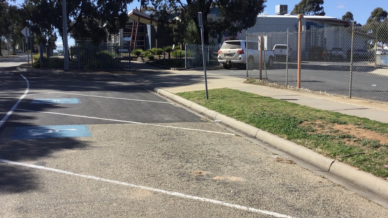 In close proximity to the centre’s main entrance is the carpark with accessible parking spaces. There is a chain link fence that serves as the carpark’s facade and a blue gate on its left where cars may enter to park. In front of the wired fence is a white pavement leading to several accessible car spaces. From this view, you will see the centre surrounded by trees.