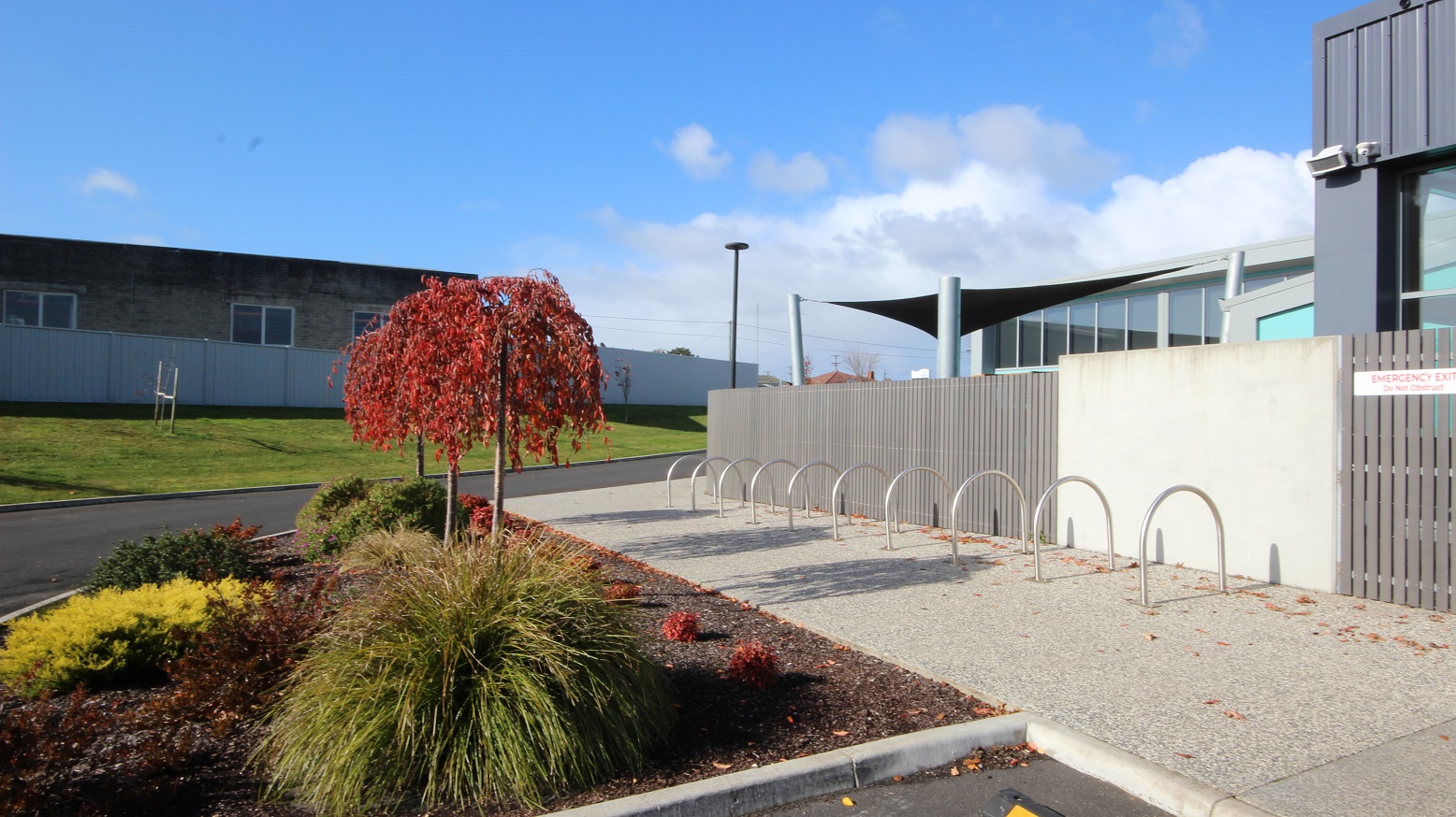 A number of inverted U-shape metal bike racks located on a rough surface of the cemented pathway near a garden allotment outside the grey fence and white wall. Inside the garden space are bushes of different types of green, yellow, green, brown, and red. Opposite the facility is a grey building with glass windows and grey, tall walls.