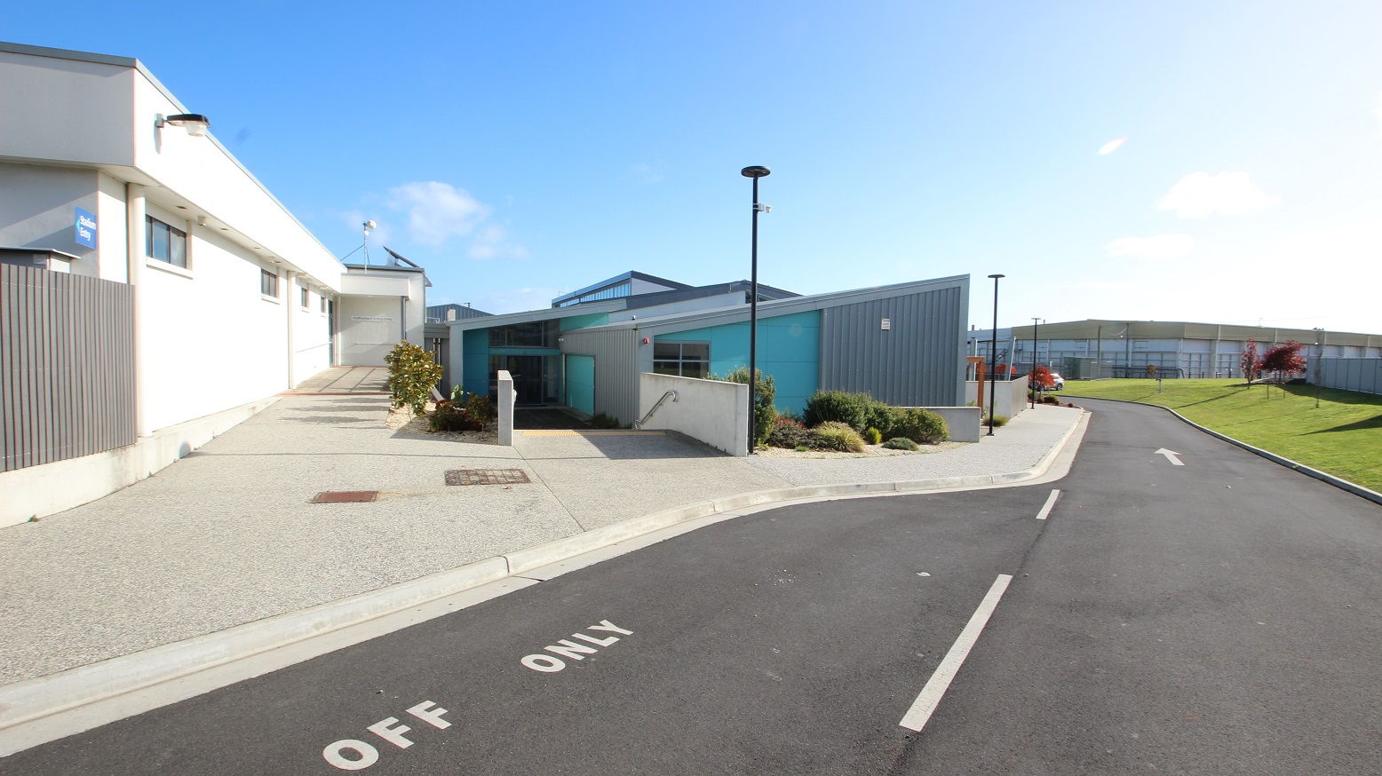 Drop-off area of SWIRL. There are two entry points to the facility.  One is through a ramp on the left where a white building with a grey fence is located while the other is through a staircase with metal handrails mounted on the hip-length white wall leading to the SWIRL, automatic, double glass door entrance. The facility is white, grey, and light green with an irregular rectangular shape with a pointed edge.
