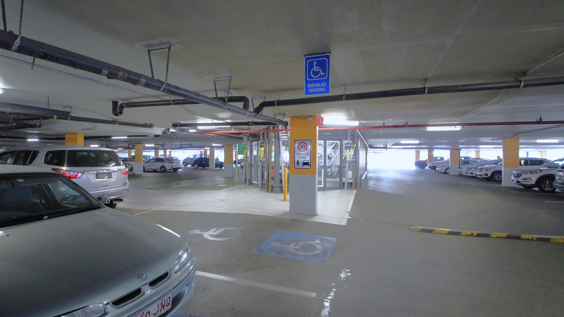 Accessible car park for persons with mobility issues in the undercover carpark at Amcal Wilsonton. The accessible car park space is close to the automatic glass sliding doors with grey metal frames in the middle of the dedicated carpark. It can be distinguished by the large blue sign on the ceiling for Disabled Parking and the white logo over a blue background. On the grey and yellow column next to the accessible car park space is a Shared Zone sign with a max speed of 10 kilometers per hour.