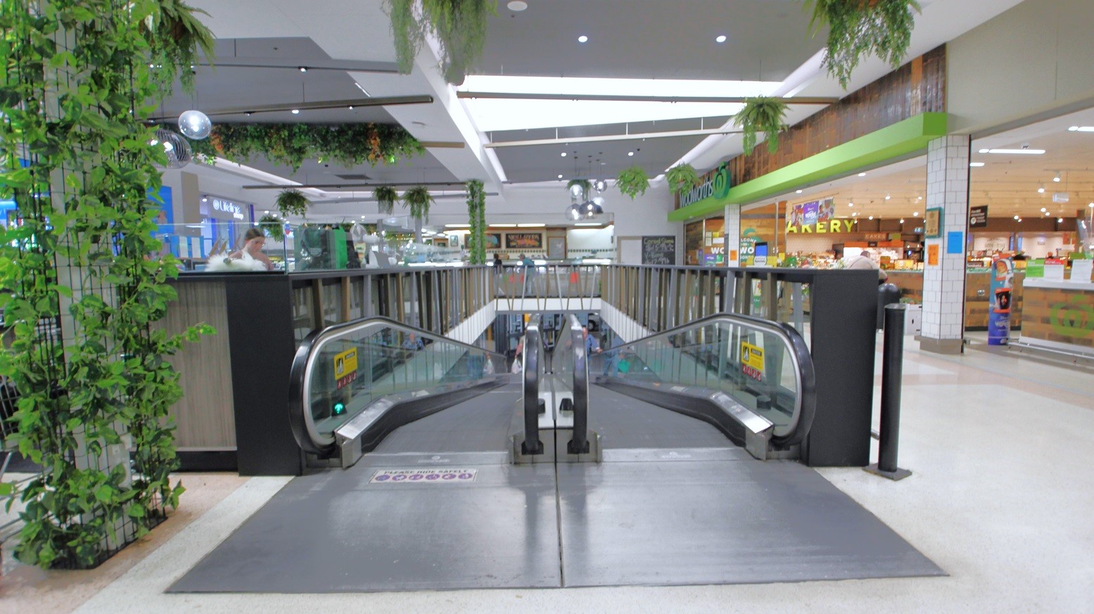 Two escalators from the undercover carpark of Bridge Street with 8000 free car parking spaces. On the left side is a column enclosed by a black screen that has green vines. Covering the other specialty retailer sections is a dark wooden handrail with light blue, brown, and grey diagonal walls. Hanging on the ceiling are green plant pots.